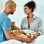 Een man en vrouw die "De liefdestaal van dienstbaarheid" tentoonstellen door een dienblad met eten in bed vast te houden.