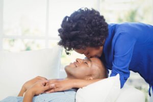 Liefde 1 Een vrouw leunt voorover en kust een man op zijn voorhoofd terwijl hij op een bank ligt, beiden omringd door witte kussens in een lichte kamer-een teder moment dat de perfecte Valentijnscadeau voor Hem vastlegt.