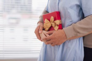 Twee mensen staan dicht bij elkaar; de een houdt een rode cadeaudoos met een gouden lint vast - een ideaal Valentijnscadeau voor Hem - terwijl de ander hen van achteren omhelst, met beide handen rustend op het ingepakte cadeau.