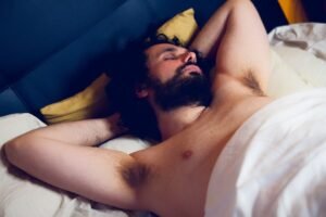 Een man met een baard ligt in bed met zijn armen achter zijn hoofd, ogen gesloten en gedeeltelijk bedekt door een witte deken.