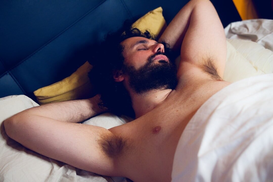 Een man met een baard ligt in bed met zijn armen achter zijn hoofd, ogen gesloten en gedeeltelijk bedekt door een witte deken.