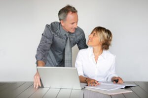 Een man staat naast een vrouw die aan een bureau zit met een open laptop en documenten; ze kijken elkaar aan en glimlachen.