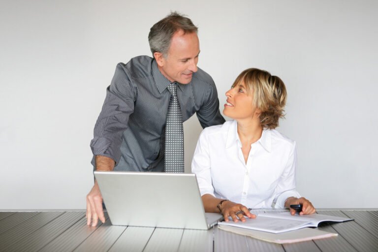 Een man staat naast een vrouw die aan een bureau zit met een open laptop en documenten; ze kijken elkaar aan en glimlachen.